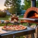Outdoor wood-fired pizza oven baking a fresh pepperoni pizza on a sunny backyard patio, with a picnic table and food in the background.